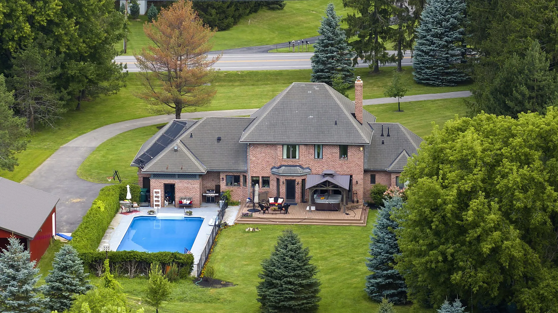 Aerial view of an in-ground pool beside a brick home and mature trees